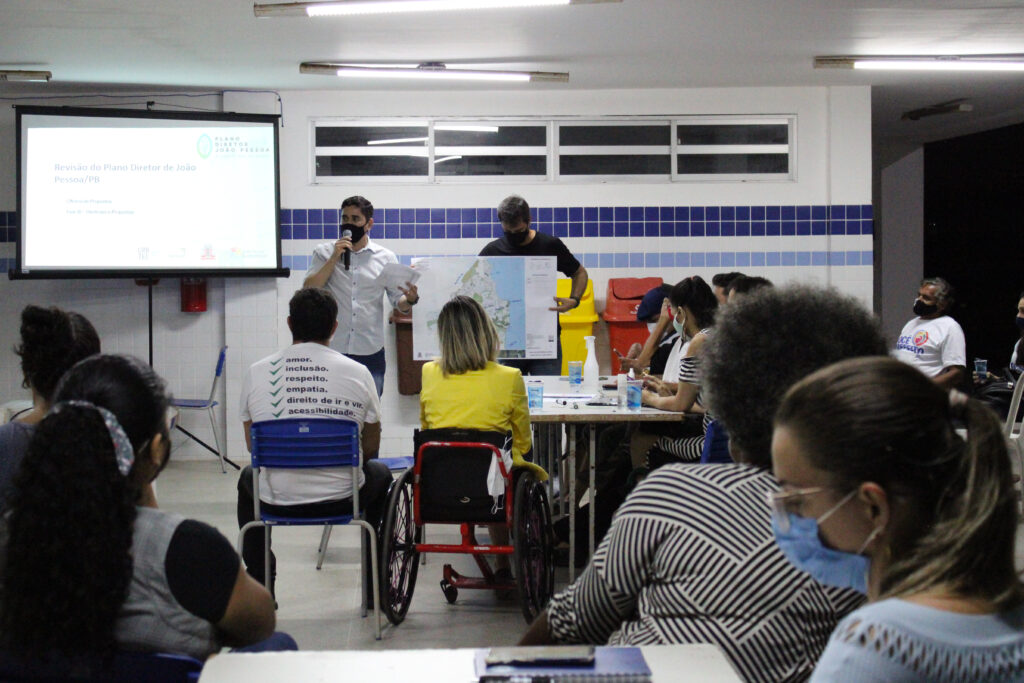 Pessoas sentadas assistindo dois participantes apresentando suas propostas.