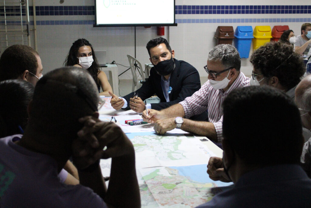 Participantes sentados discutindo propostas, com mapas sobre a mesa